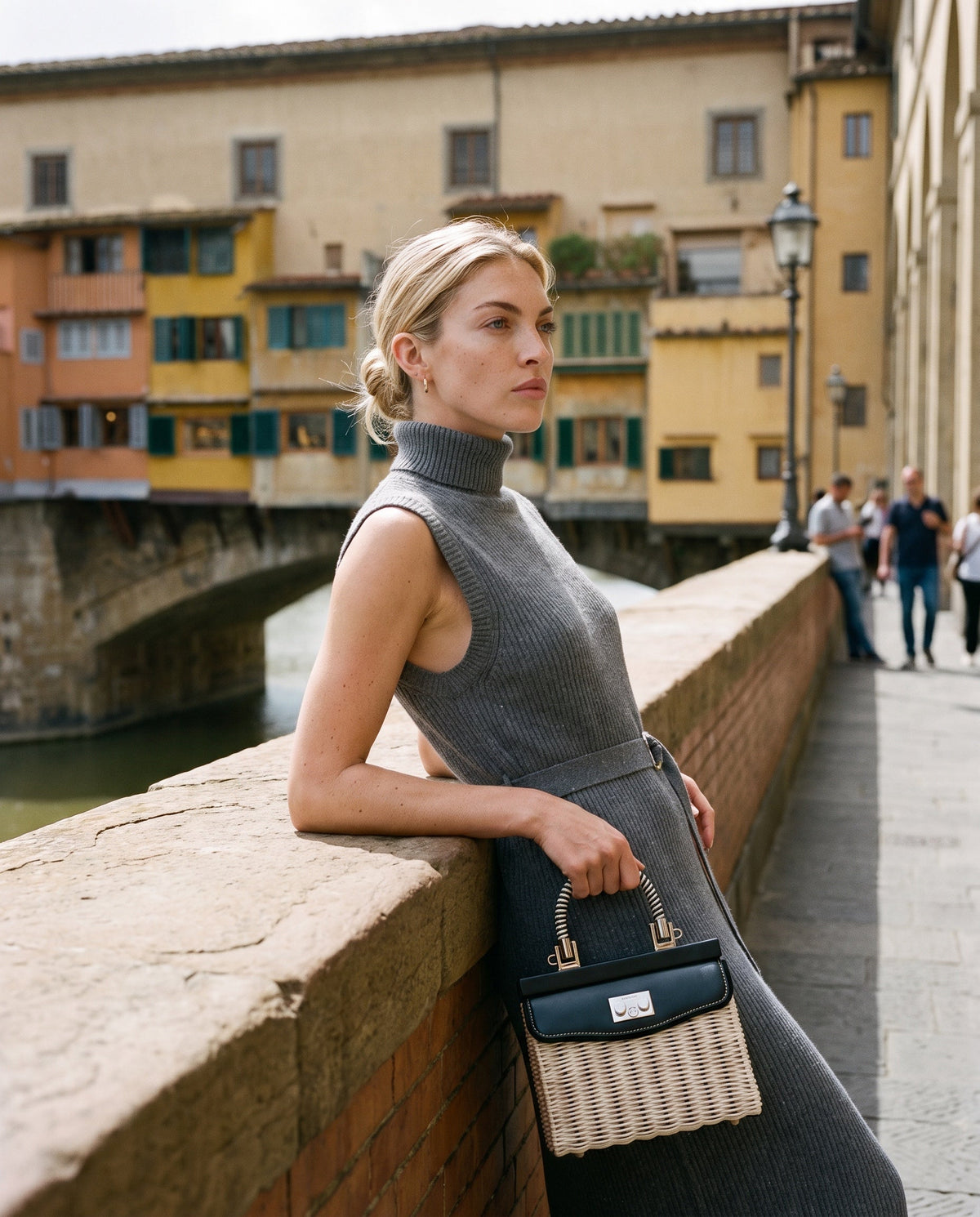 Model in a grey sleeveless rib-knit midi dress with a wicker top-handle bag by the Arno, opposite the Ponte Vecchio